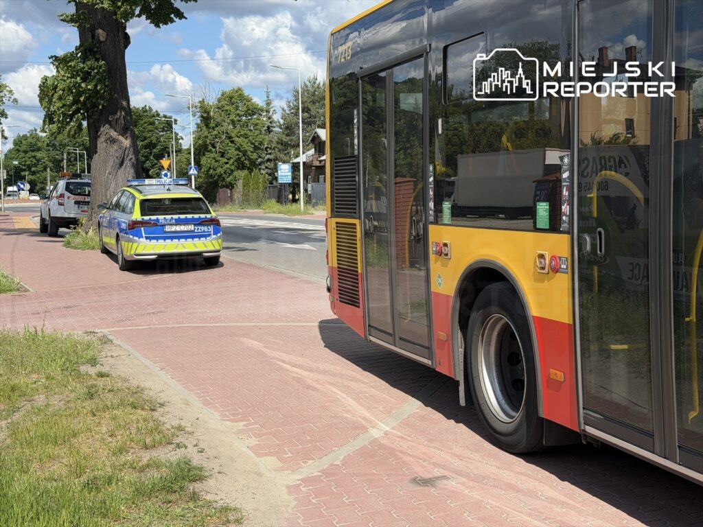 Policyjny radiowóz zaparkowany na chodniku obok przystanku autobusowego, obok czerwono-żółtego autobusu.