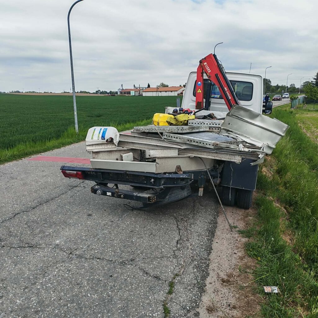 Na drodze stoi przyczepa samochodowa załadowana metalowymi elementami i narzędziami, w tle pola i zabudowania.