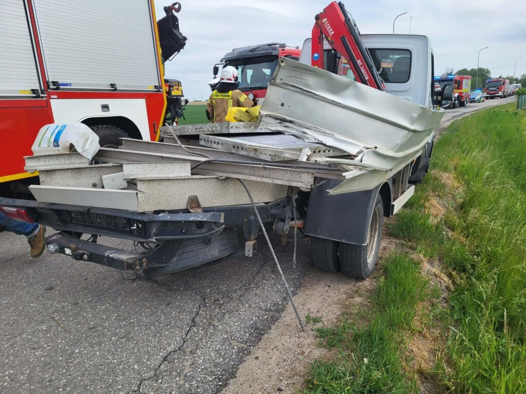 Uszkodzony samochód dostawczy z rozbitej skrzynią ładunkową stoi na poboczu drogi, obok interweniujący strażacy.