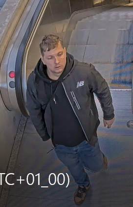 Mężczyzna w ciemnej kurtce i dżinsach schodzi po ruchomych schodach w przestronnym pomieszczeniu.