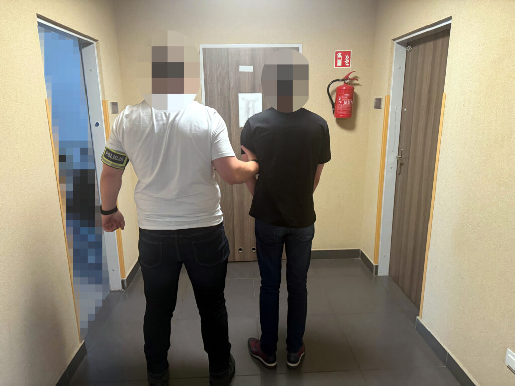 Funkcjonariusz w białej koszulce z napisem "POLICJA" prowadzi osobę w czarnej koszulce korytarzem w budynku.