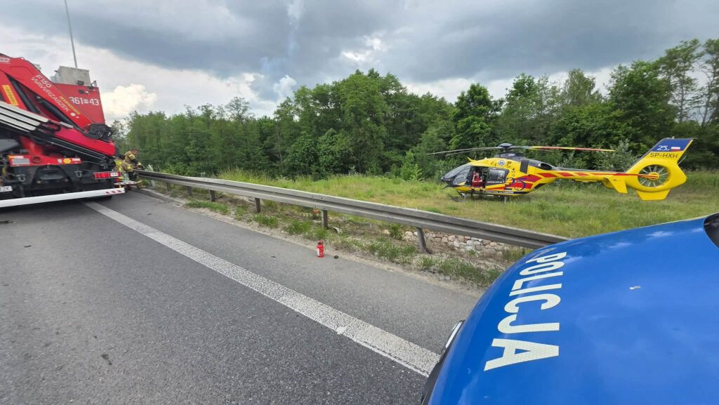 Strażak przy barierce na drodze, obok czerwona ciężarówka ratunkowa i żółty helikopter ratunkowy w tle.