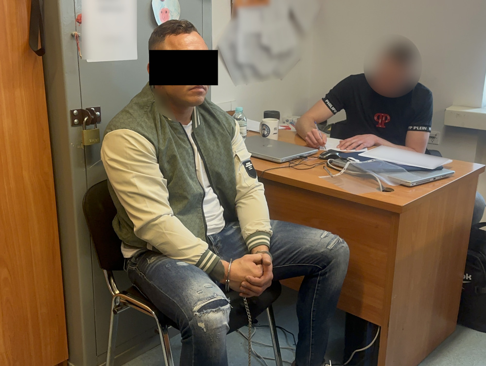 Mężczyzna w sportowej kurtce siedzi na krześle, podczas gdy osoba przy biurku pisze notatki. W tle widoczna szafa.