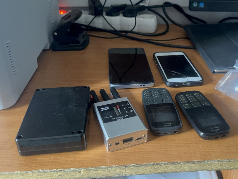Na drewnianym biurku leżą różne urządzenia elektroniczne: dwa telefony komórkowe, tablet oraz detektor RF.
