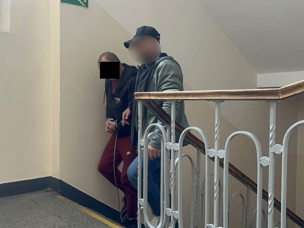 Dwóch mężczyzn schodzących po schodach, jeden z nich w kajdankach, w budynku o jasnych ścianach.