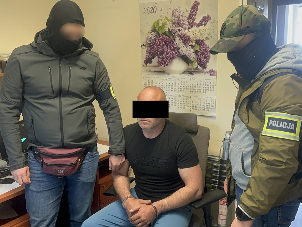 Mężczyzna siedzący na krześle, otoczony przez dwóch funkcjonariuszy w maskach, w biurze z kalendarzem na ścianie.