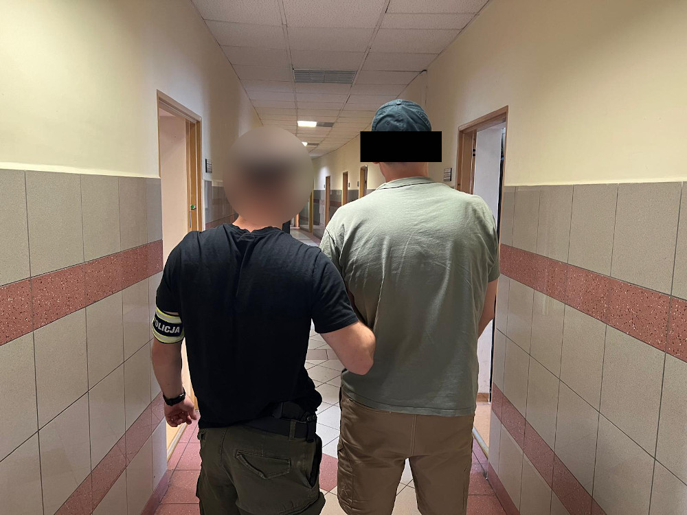 Funkcjonariusz w mundurze prowadzi mężczyznę korytarzem w budynku administracyjnym.