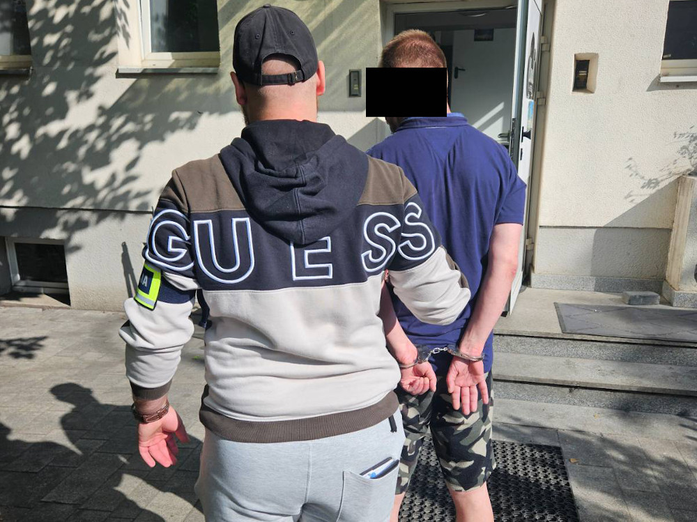 Mężczyzna w szarej bluzie zakuwający w kajdanki drugiego mężczyznę na schodach budynku.