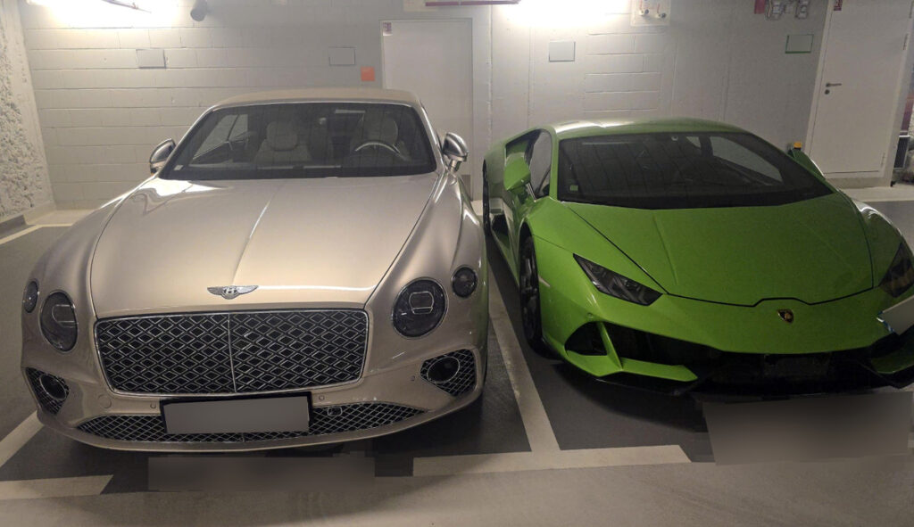 Dwa luksusowe samochody zaparkowane obok siebie w podziemnym garażu: srebrny Bentley i zielony Lamborghini.