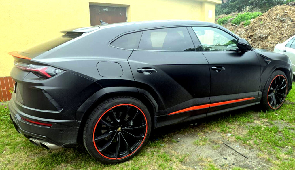 Czarny SUV z matowym wykończeniem stoi na trawie obok żółtej ściany budynku, z wyraźnymi czerwonymi detalami na felgach.