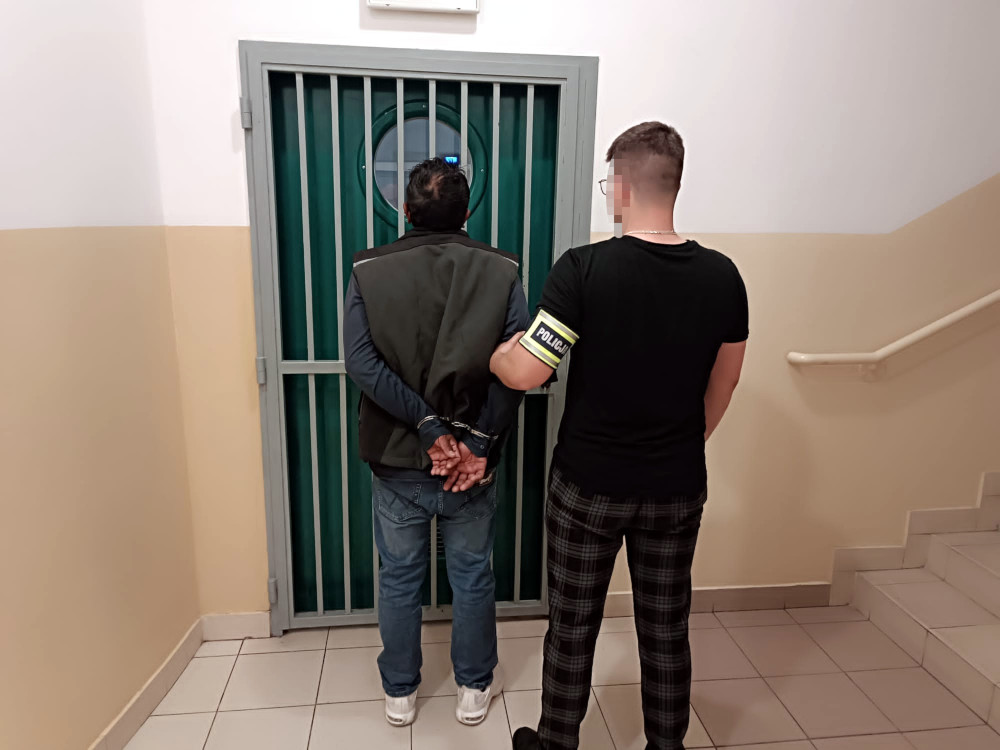 Mężczyzna w kajdankach stoi tyłem przy drzwiach z kratami, a policjant w koszulce z napisem "POLICJA" obserwuje sytuację.