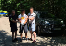 Mężczyzna z opatrunkiem na ręce jest prowadzony przez dwóch funkcjonariuszy w lesistym otoczeniu, obok stoi samochód.
