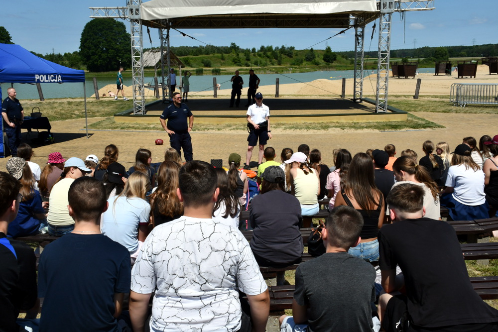 Grupa młodzieży siedzi na ławkach, słuchając policjantów przemawiających na scenie w plenerze.