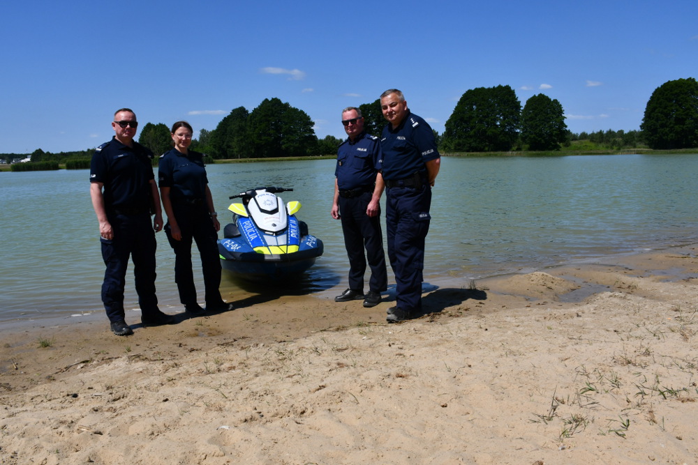 Czterech funkcjonariuszy w mundurach stoi na plaży przy jeziorze obok skuteru wodnego.