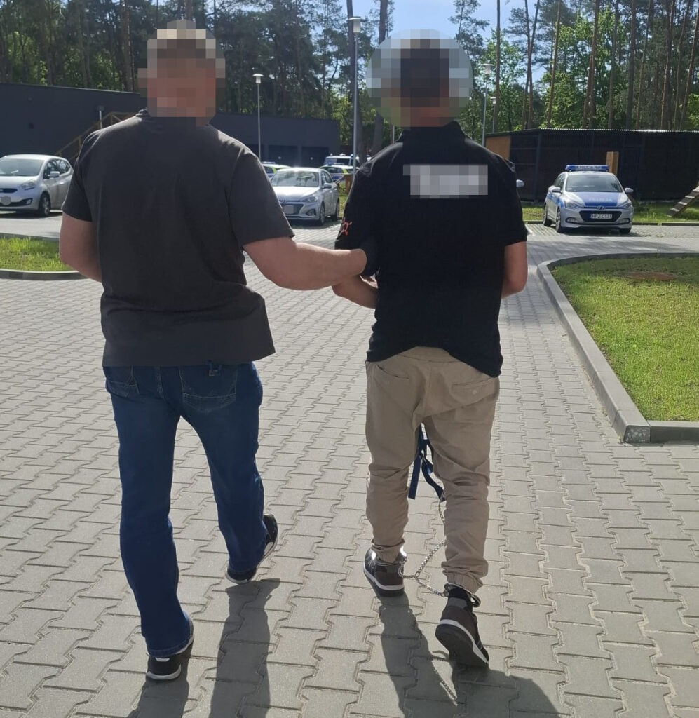Mężczyzna w garniturze prowadzi osobę w spodniach beżowych na uwięzi, idąc po chodniku w otoczeniu zaparkowanych samochodów.