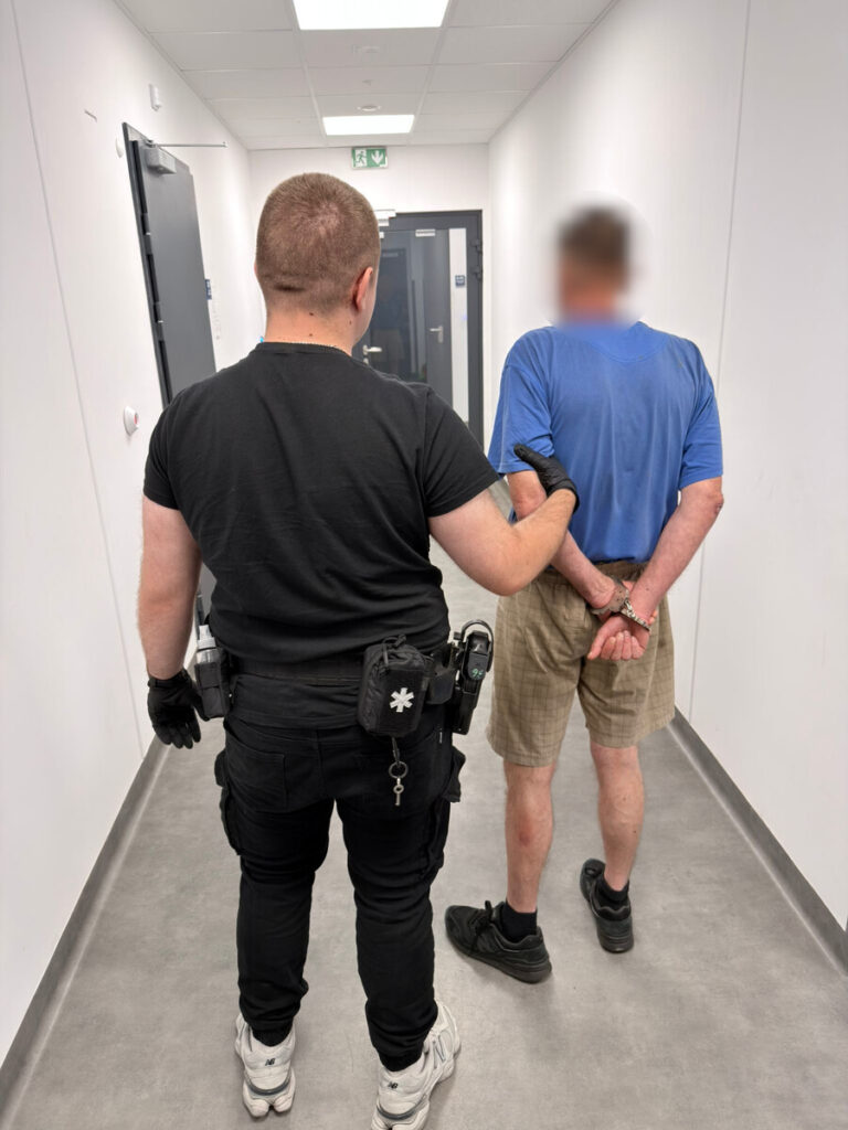 Funkcjonariusz policji prowadzi zatrzymanego mężczyznę w niebieskiej koszulce korytarzem budynku.