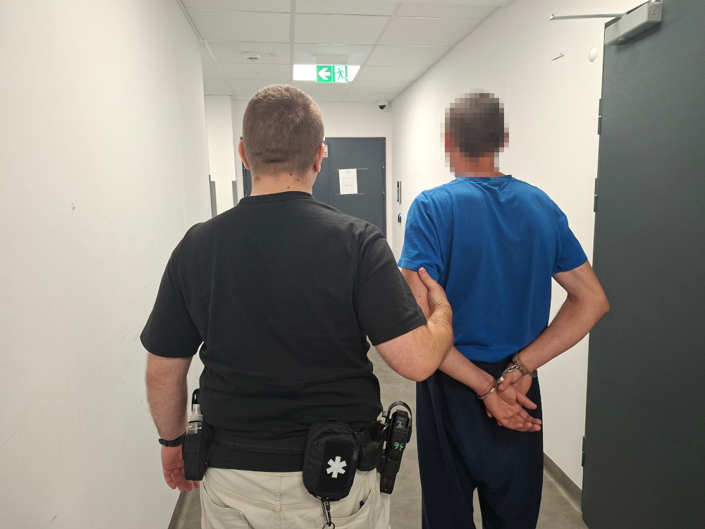 Funkcjonariusz policji prowadzi mężczyznę w niebieskiej koszulce z rękami związanymi z tyłu korytarzem w budynku.