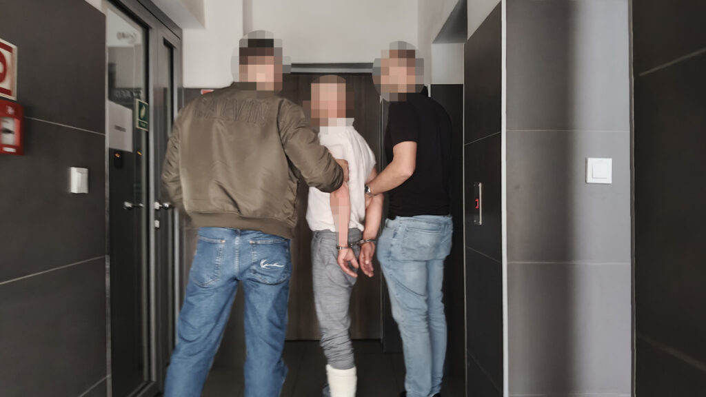 Mężczyzna w kajdankach prowadzony przez dwóch funkcjonariuszy w nowoczesnym korytarzu budynku.