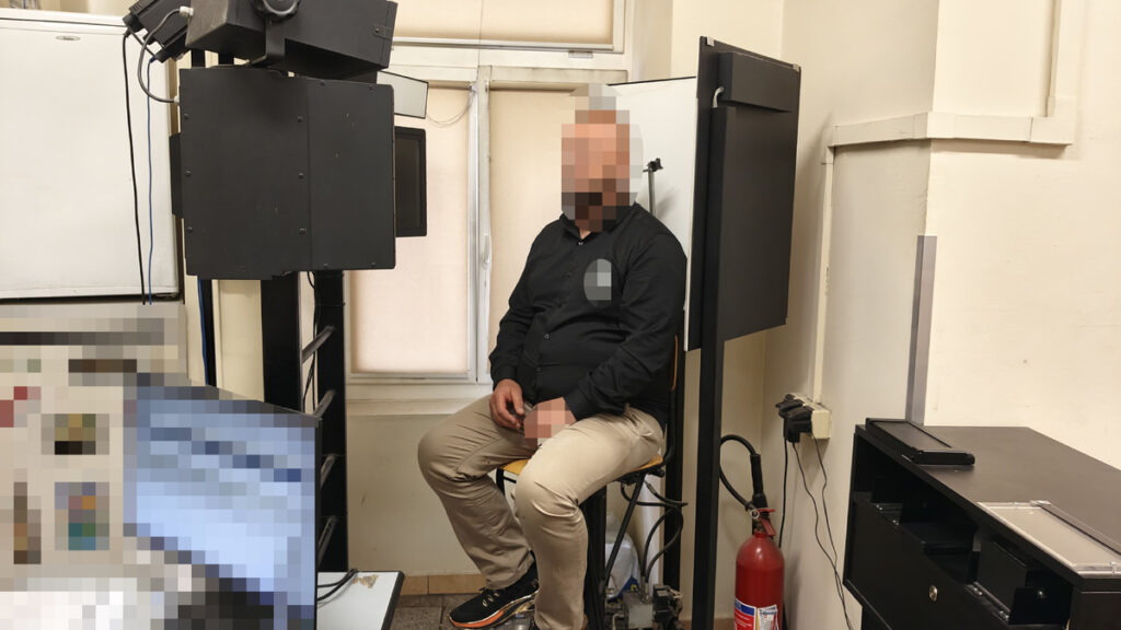 Mężczyzna w czarnej koszuli siedzi na krześle przed sprzętem fotograficznym w jasnym pomieszczeniu.