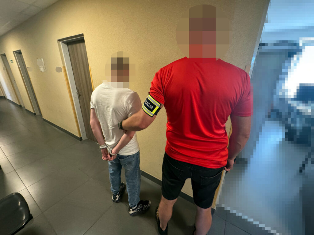 Mężczyzna w białej koszulce z rękami związanymi z tyłu stoi obok funkcjonariusza w czerwonym ubraniu z opaską "POLICJA" w korytarzu.