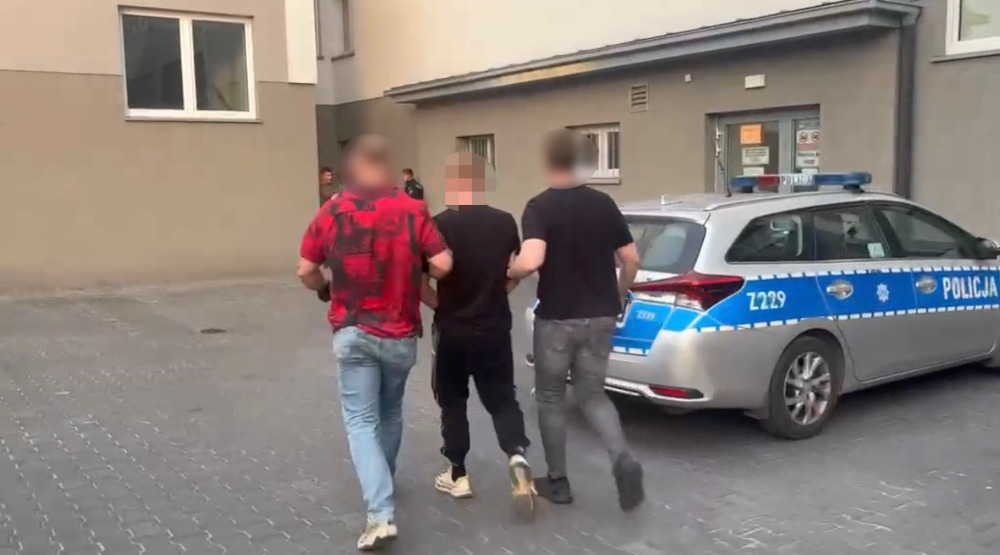 Mężczyzna w czarnym ubraniu prowadzony przez dwóch policjantów w pobliżu radiowozu na parkingu.