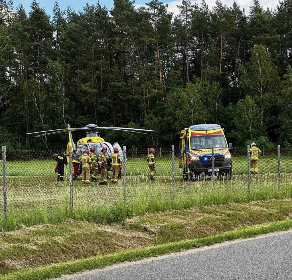 Grupa ratowników w mundurach podchodzi do śmigłowca ratunkowego na tle lasu, obok stoi ambulans.