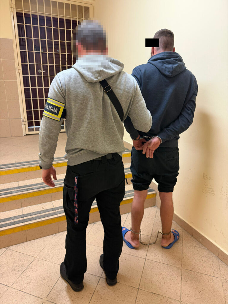 Funkcjonariusz policji prowadzi mężczyznę w kajdankach w budynku policyjnym.