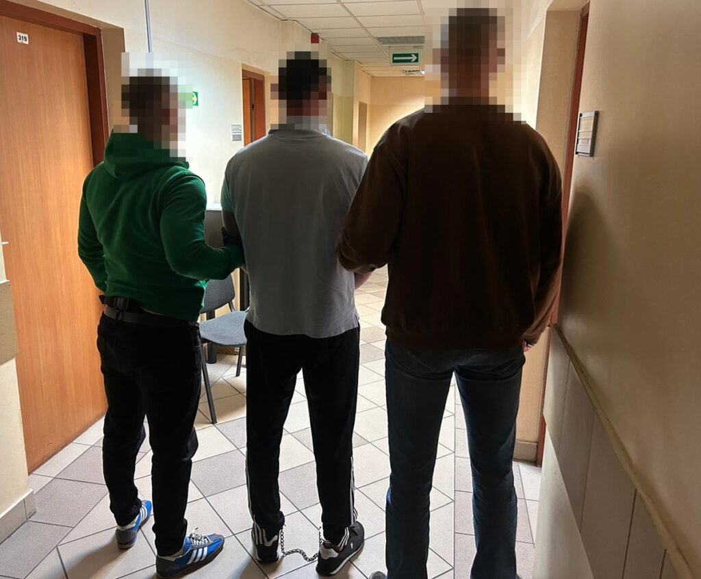 Trzej mężczyźni stoją w korytarzu budynku, jeden z nich ma założone kajdanki, a pozostali trzymają go za ramiona.