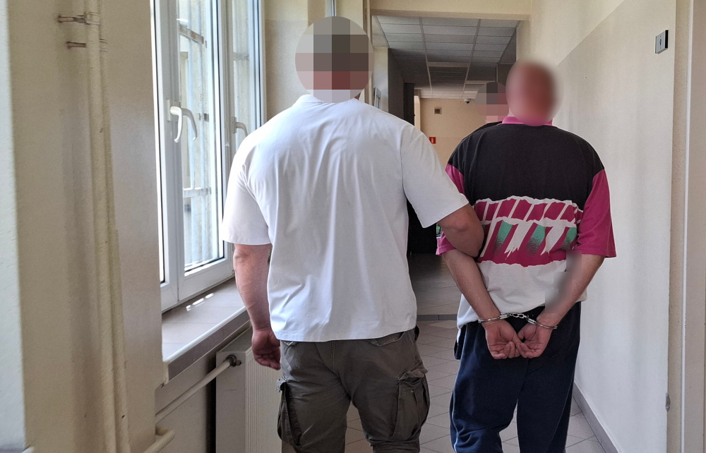 Mężczyzna w białej koszulce prowadzi aresztowanego w kajdankach korytarzem budynku.