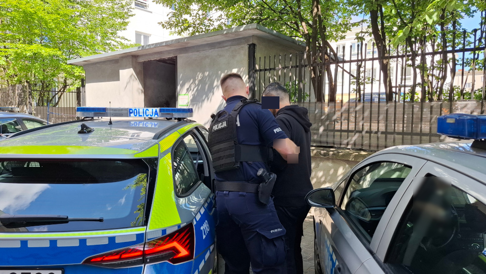 Policjant w kamizelce ochronnej zatrzymuje mężczyznę w czarnej bluzie obok radiowozu na tle ogrodzenia.