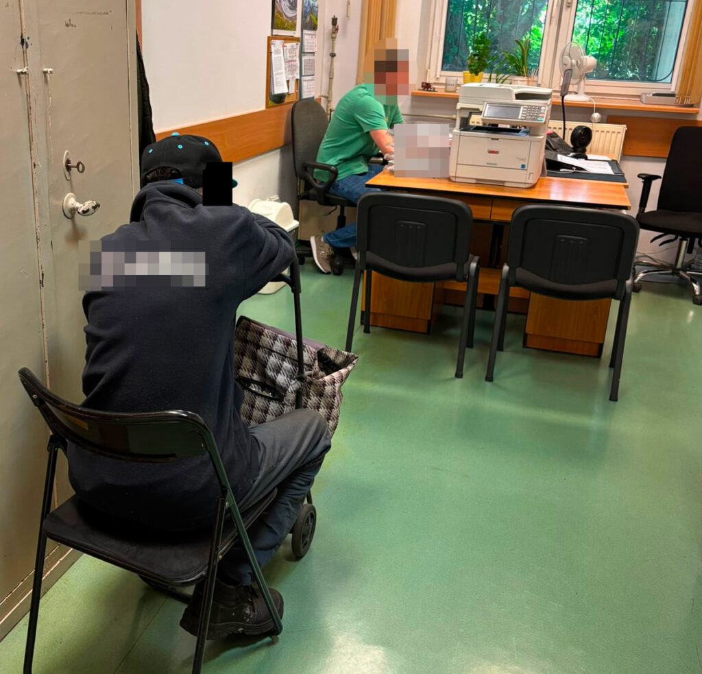 Mężczyzna w ciemnej odzieży siedzący na krześle z torbą na kółkach, obserwujący rozmowę przy biurku.