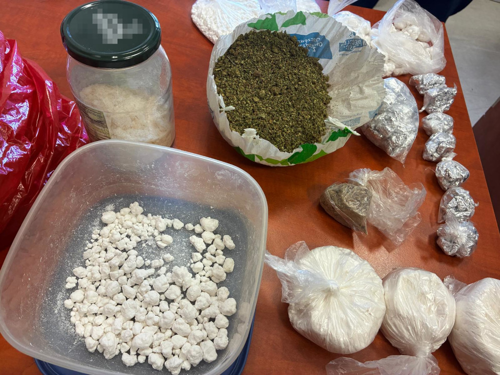 Na stole znajdują się różne opakowania z substancjami, w tym biały proszek, zielona marihuana oraz foliowe torebki.