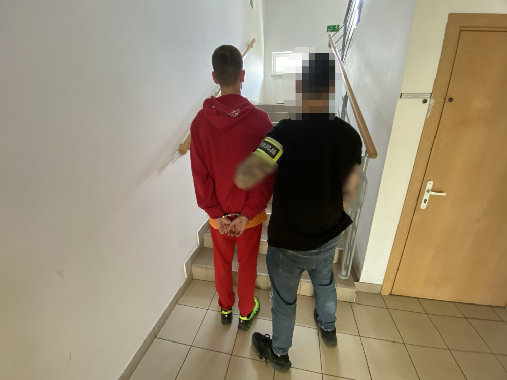 Młody mężczyzna w czerwonym dresie prowadzony przez funkcjonariusza policji na schodach w budynku.