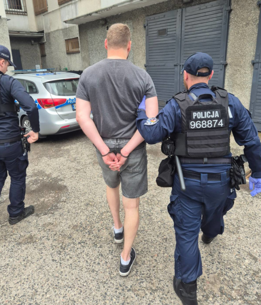 Mężczyzna w szortach prowadzony przez policjantów w mundurach w pobliżu zaparkowanego radiowozu.
