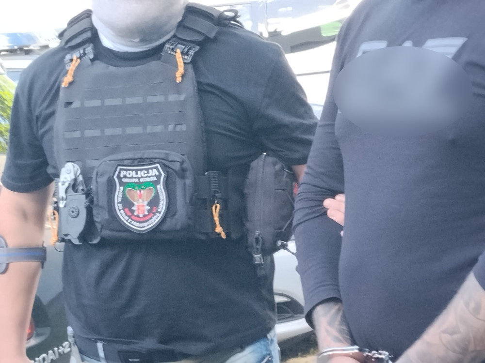 Mężczyzna w kamizelce kuloodpornej z logo policji stoi obok mężczyzny z rękami skrępowanymi w kajdankach.