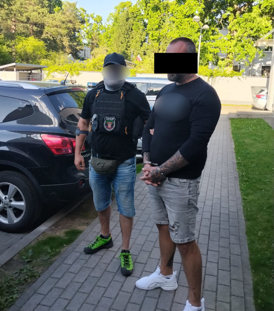 Funkcjonariusz w kamizelce taktycznej stoi obok mężczyzny w kajdankach, na chodniku w otoczeniu zaparkowanych samochodów.