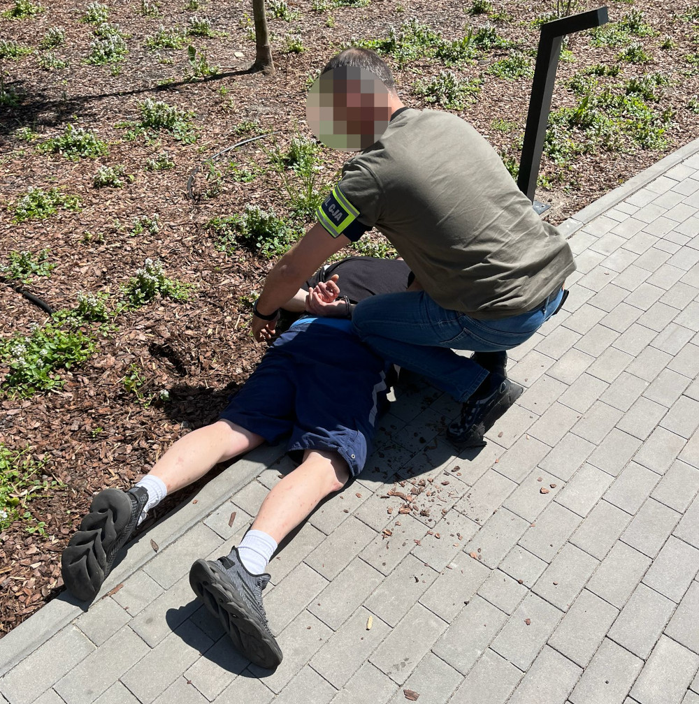 Mężczyzna w mundurze zatrzymuje leżącego na ziemi mężczyznę w parku, otoczonym przez zieleń.