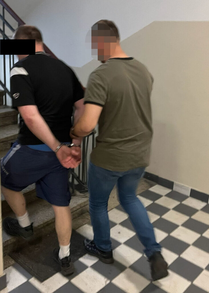 Mężczyzna w kajdankach prowadzony przez funkcjonariusza po schodach w budynku z wyłożoną szachownicą podłogową.