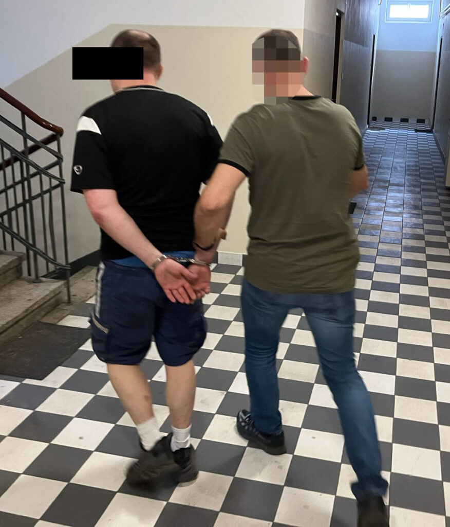 Mężczyzna w kajdankach prowadzony przez innego mężczyznę w korytarzu z szachownicową podłogą.