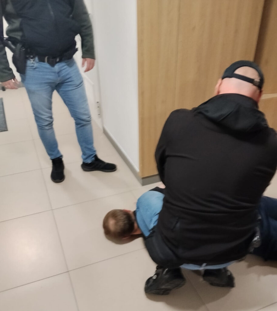 Mężczyzna leży na podłodze, a dwóch funkcjonariuszy policji trzyma go w areszcie w nowoczesnym wnętrzu budynku.