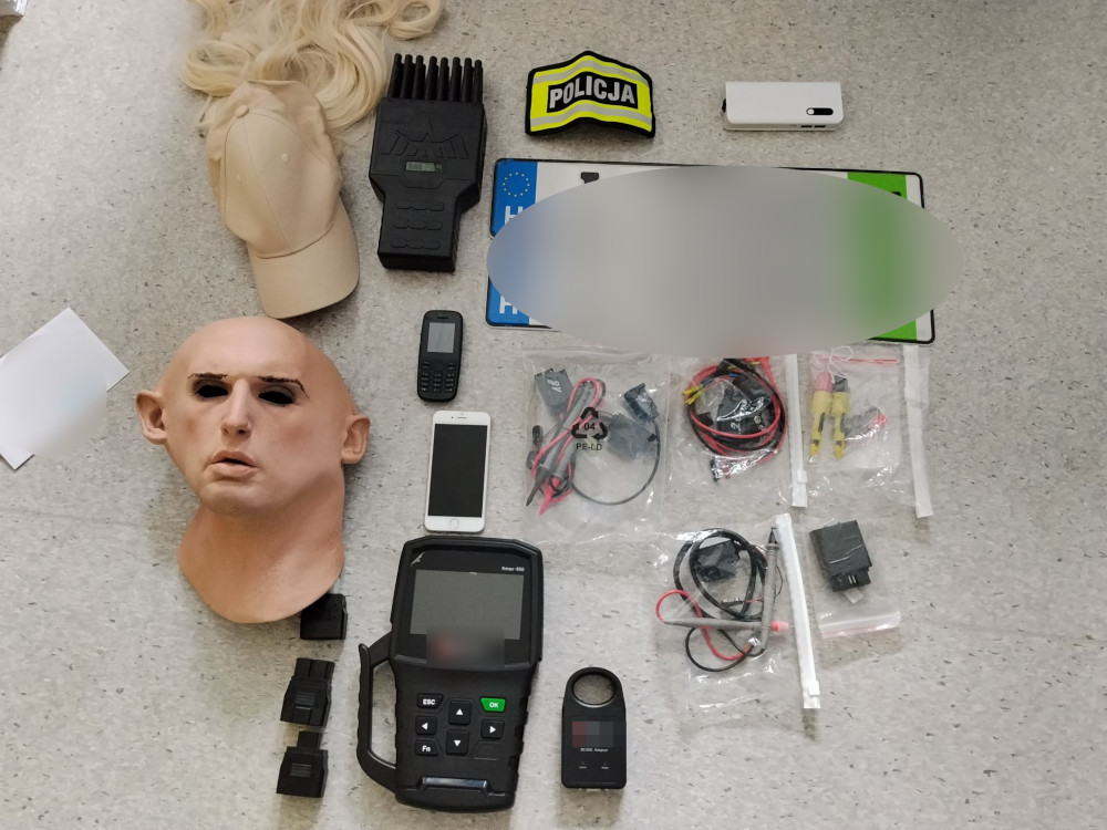 Na blacie leżą różne przedmioty, w tym maska głowy, telefon komórkowy, urządzenie pomiarowe oraz akcesoria elektroniczne. W tle widoczna czapka i naszywka z napisem "POLICJA".