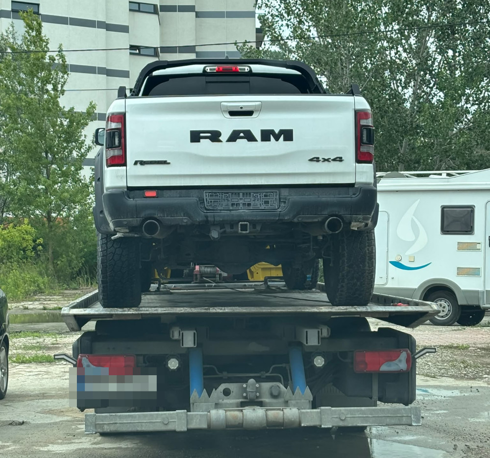 Biały pickup RAM 4x4 załadowany na lawetę, stojący na placu z zielenią w tle.