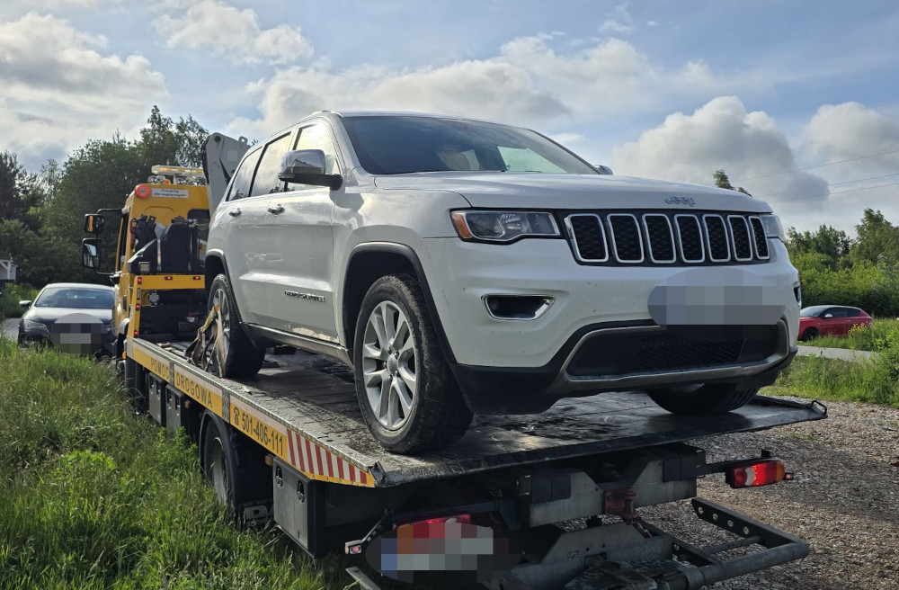 Biały SUV marki Jeep załadowany na lawetę w otoczeniu zieleni i innych pojazdów na drodze.