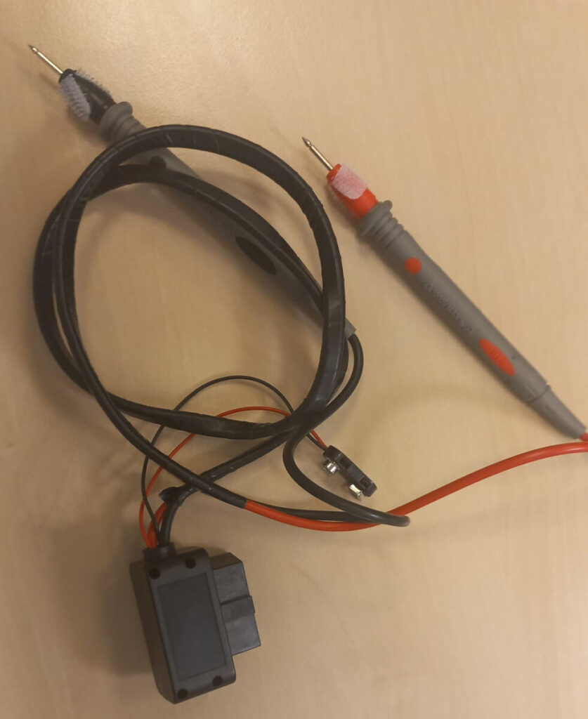 Zestaw przewodów pomiarowych z końcówkami oraz modułem elektronicznym leżący na drewnianym biurku.
