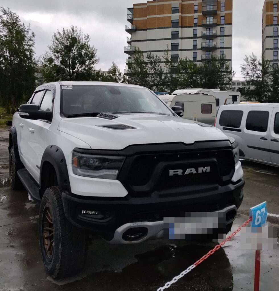 Biały pick-up RAM zaparkowany na terenie parkingu, w tle nowoczesne budynki mieszkalne i zielone drzewa.