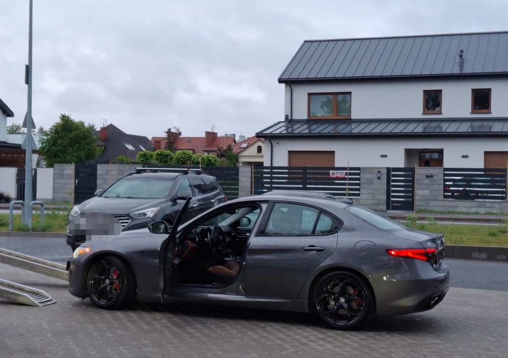 Szary samochód osobowy z otwartymi drzwiami stoi na parkingu przed nowoczesnym domem z przeszkloną fasadą.