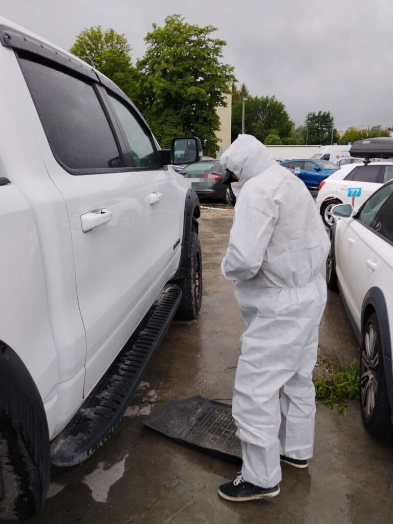 Mężczyzna w białym kombinezonie ochronnym sprawdza pojazd w deszczowej aurze na parkingu.
