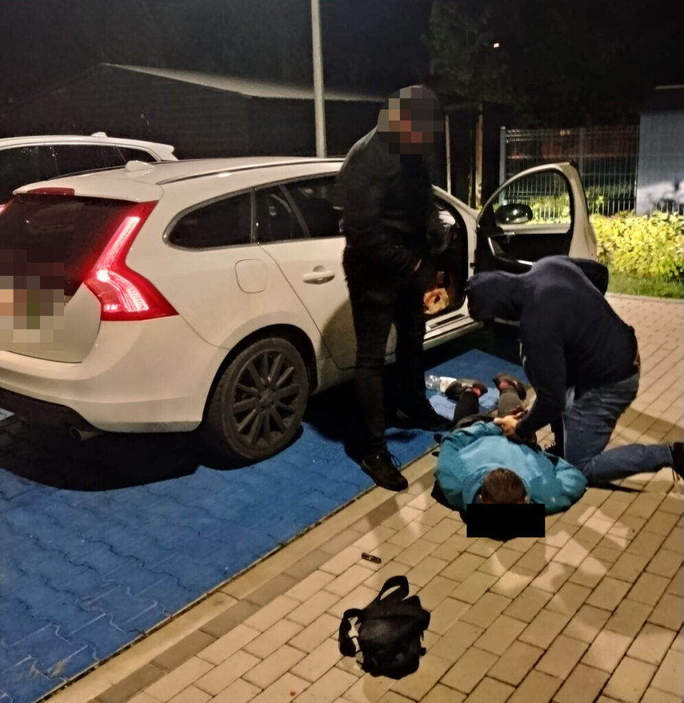 Dwaj mężczyźni w ciemnych ubraniach przeszukują leżącego na ziemi mężczyznę w niebieskiej kurtce obok białego samochodu.