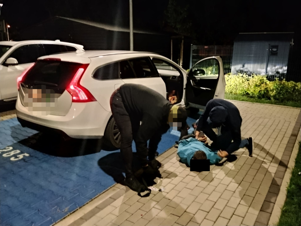 Dwaj mężczyźni udzielają pomocy osobie leżącej na ziemi obok białego samochodu w nocy.