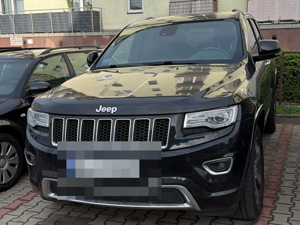 Czarny SUV marki Jeep zaparkowany na chodniku w miejskim otoczeniu, obok innych samochodów.